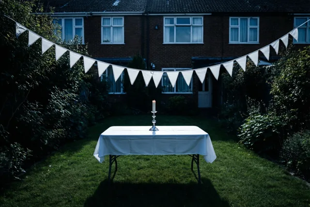 Een witte tafel met een wit kleed en één communiekaars staat op het gras achter een rijhuis, met witte driehoekige vlaggetjes en groen in gedempt licht.