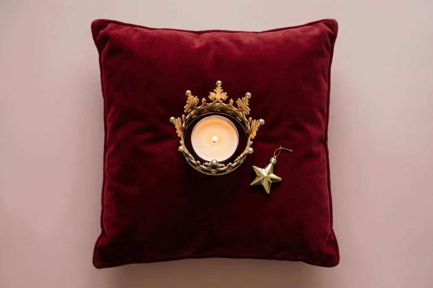 Een brandende kaars in een gouden kroonvormige houder en een verguld sterornament staan op een dieprood fluwelen kussen tegen een effen lichtroze achtergrond.