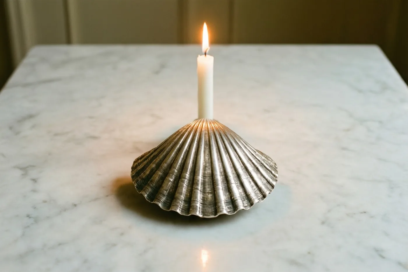 Een slanke witte kaars brandt in een zilveren kandelaar in de vorm van een doopschelp op wit marmer.