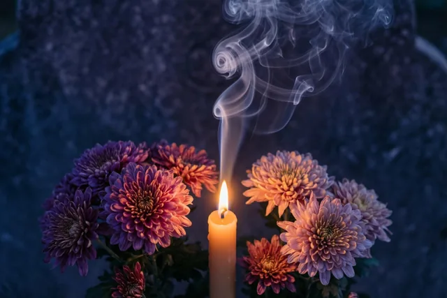 Een brandende kaars met een dun sliertje rook aan de lont staat naast paarse, roze en oranje chrysanten tegen een donkere achtergrond met textuur.