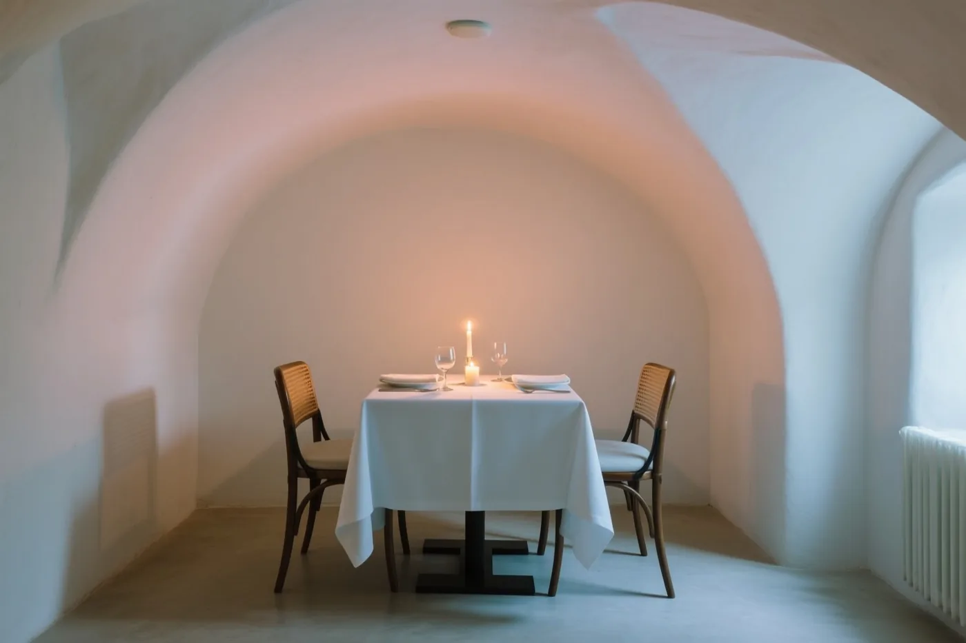 Een tafel met een wit tafelkleed en houten stoelen staat voor twee personen gedekt onder een wit gewelf in een schemerig kelderrestaurant, verlicht door één kaars.