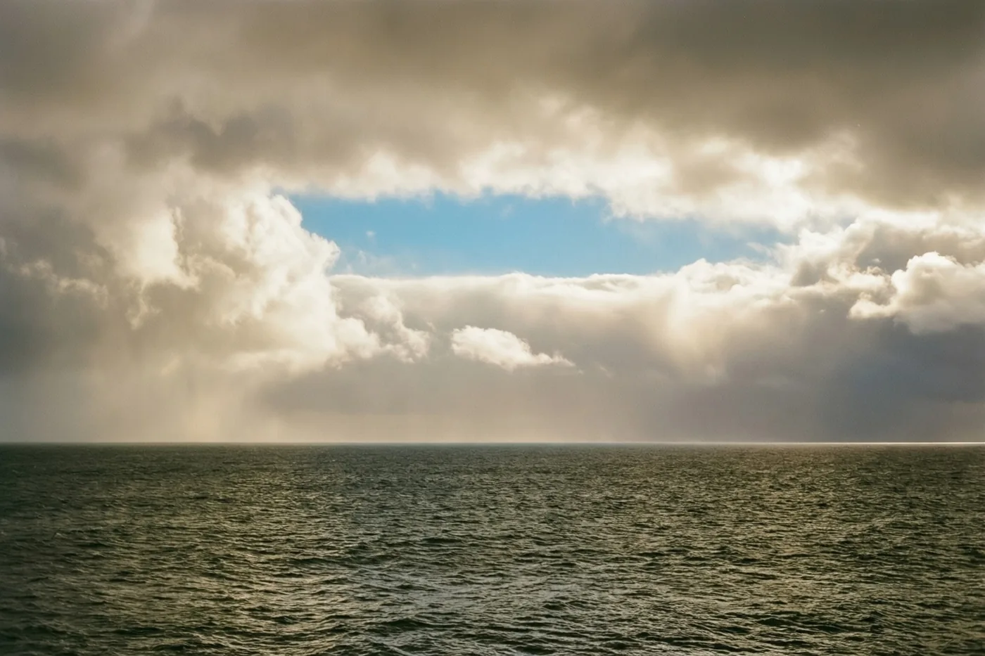 Een kalme zee ligt onder openbrekende wolken met een smalle strook blauwe lucht, in gedempte groene en grijze tinten.