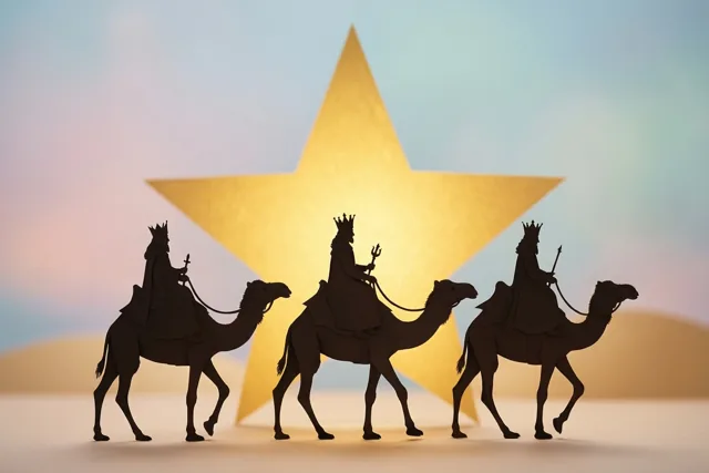 Silhouetten van drie koningen op kamelen trekken achter elkaar langs een grote gouden ster, met op de achtergrond een lucht in zacht verlopende warme tinten.