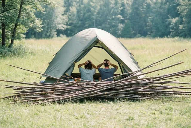 Twee mensen zitten met hun rug naar de camera in een ingestorte tent tussen houten stokken op een zonnig grasveld met bomen op de achtergrond.