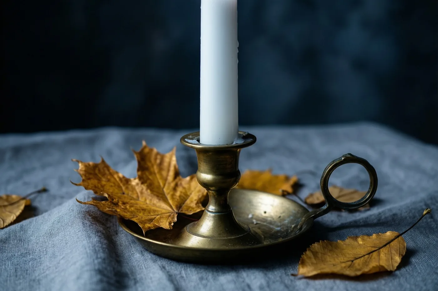 Een hoge witte kaars in een messing kandelaar staat op grijs linnen tussen gouden herfstbladeren, tegen een donkerblauwe achtergrond.