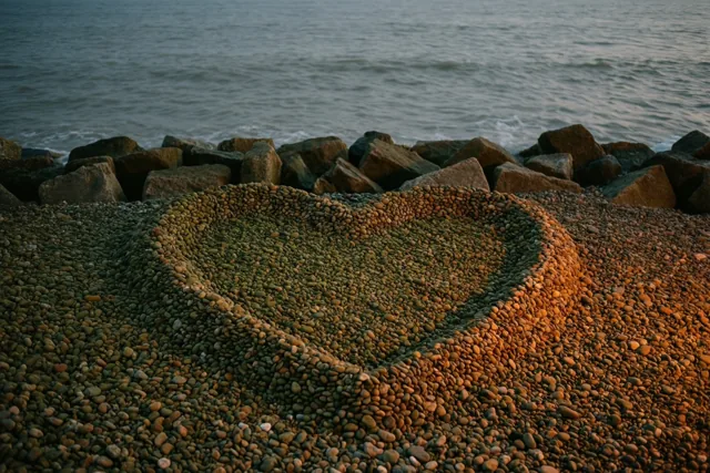 Een hart van keien ligt op een kiezelstrand voor een stenen golfbreker, met een grijze zee op de achtergrond in warm goudkleurig licht.