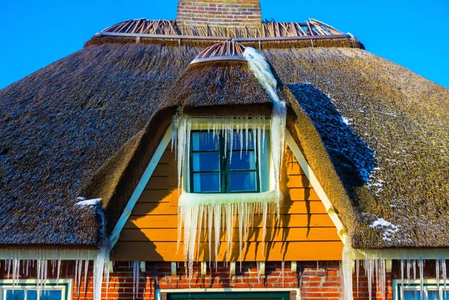 IJspegels hangen aan de dakrand en het raamkozijn van een Kempense boerderij met een rieten dak en oranje houten gevelbekleding onder een blauwe lucht.