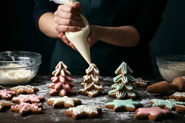 Iemand spuit wit glazuur op peperkoekkoekjes in boomvorm op een donkere houten tafel met bloemsuiker en andere versierde koekjes, tegen een donkere achtergrond.