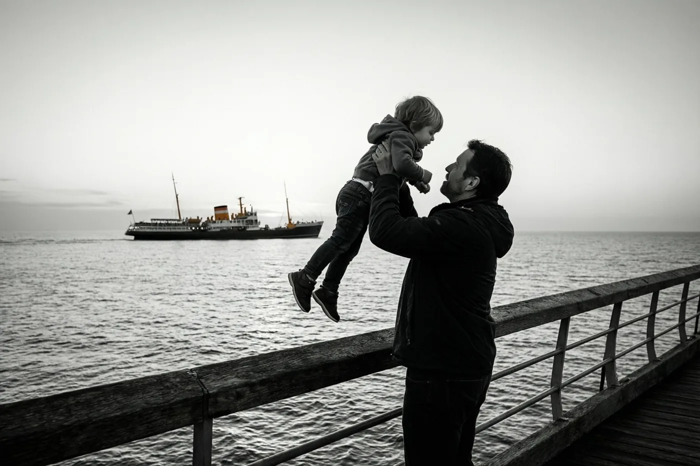 Een zwart-witfoto toont een vader die een klein kind op een houten pier in de lucht tilt, terwijl ze naar een schip in de verte op zee kijken.