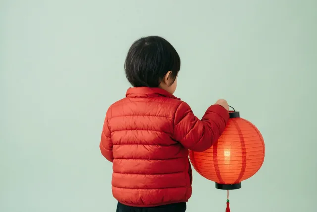 Een kind in een rode gewatteerde jas houdt een brandende rode papieren lantaarn vast, van achteren gezien tegen een effen lichtgroene achtergrond.