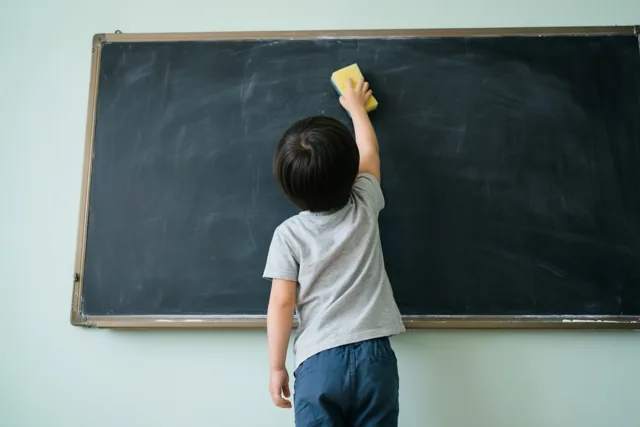Een klein kind in een grijs shirt en een blauwe broek staat op de tenen en poetst een schoolbord met een gele spons, tegen een lichtgroene muur.