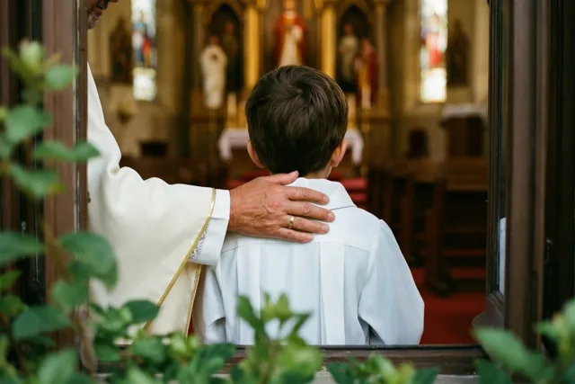 Een peter of meter staat van achteren gezien met een hand op de schouder van een kind in een wit gewaad in een warm verlichte kerk met rode loper en groen op de voorgrond.