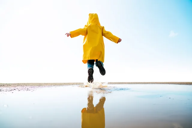 Een kind in een felgele regenjas springt met de rug naar de kijker over een plas onder een heldere hemel, met een weerspiegeling in het water.