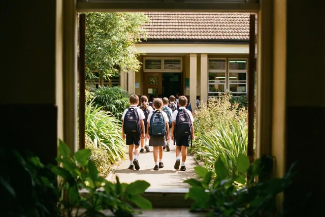 Door een deuropening is een groep schoolkinderen met rugzakken van achteren te zien op een pad naar een schoolgebouw, met groen op de voorgrond en warm zonlicht.