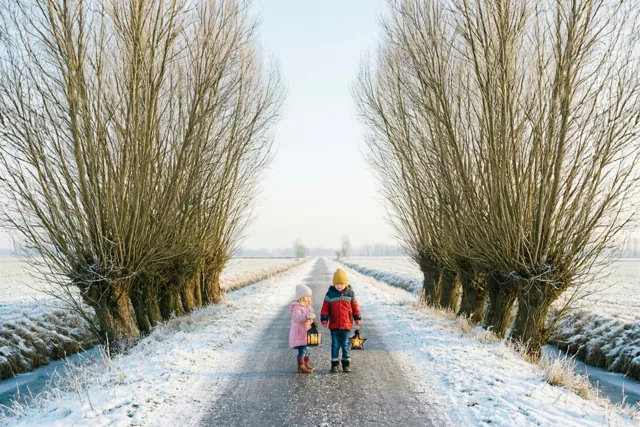 Twee kinderen lopen elk met een lichtgevende lantaarn over een lichtbesneeuwde weg tussen knotwilgen in een winterse polder, in zachte witte, grijze en bruine tinten.