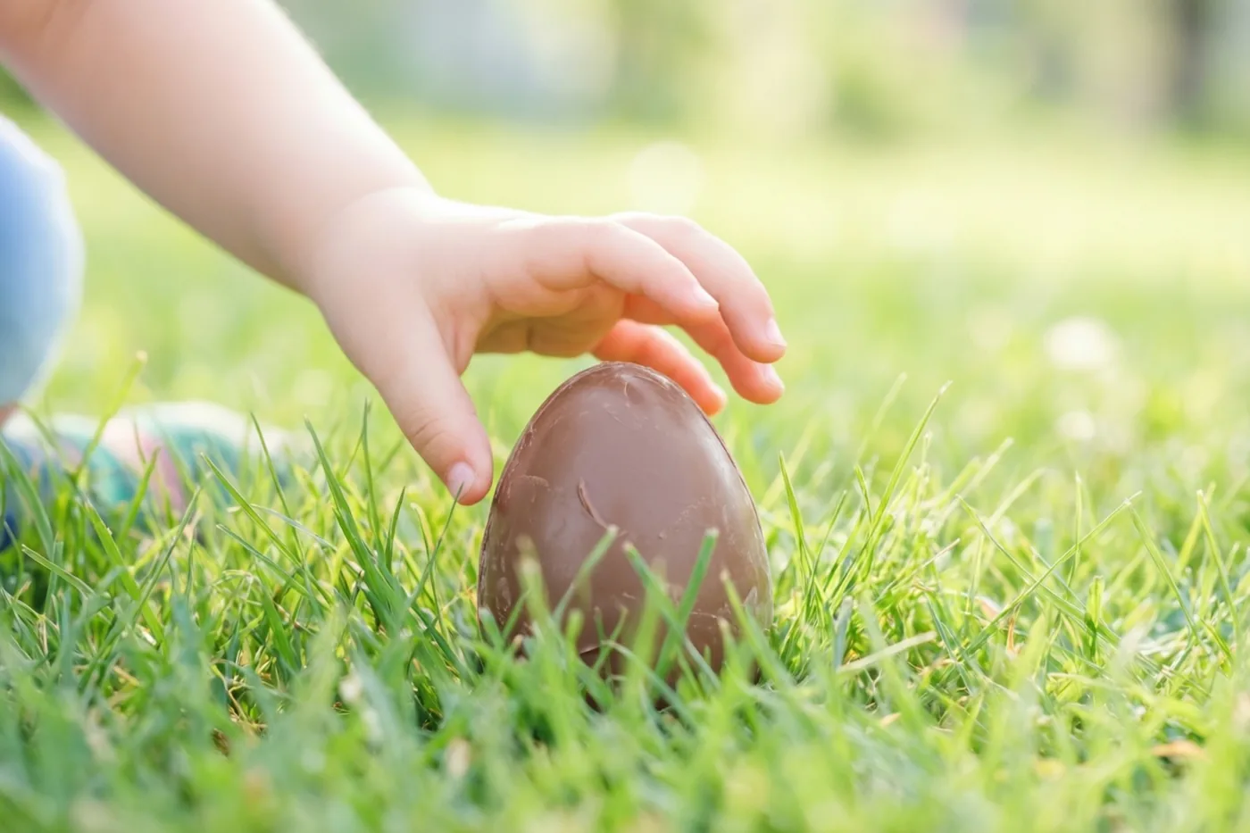 Een kinderhand reikt naar een groot chocolade-ei in felgroen gras in zacht zonlicht.