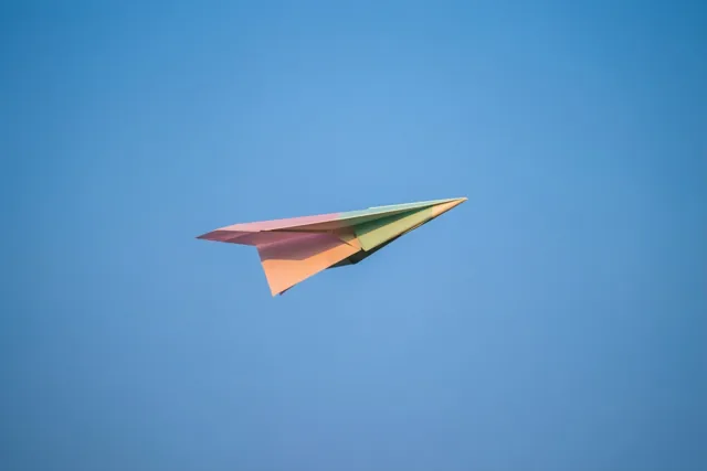 Een kleurrijk papieren vliegtuigje met blauw, roze en groen vliegt door een heldere blauwe lucht.
