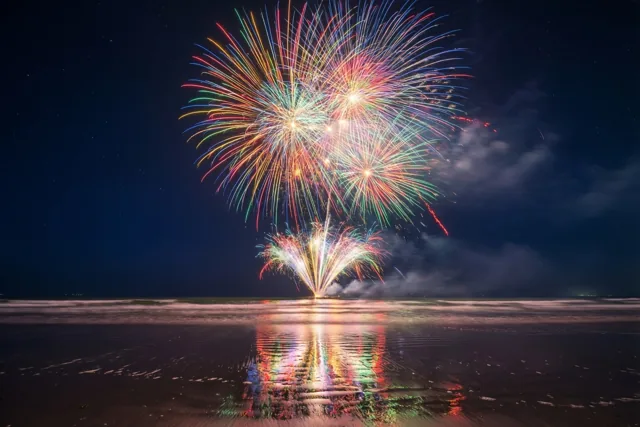 Rood, groen en blauw vuurwerk barst los boven een donker strand en weerspiegelt in het natte zand onder een diepblauwe nachtlucht.