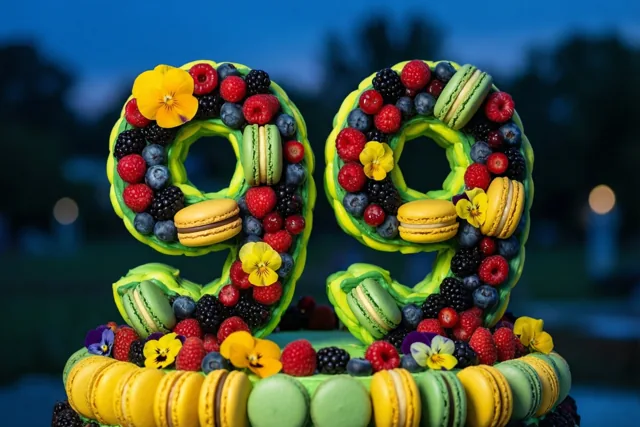 Een felgekleurde taart in de vorm van 99 staat buiten in de schemering, versierd met bessen, macarons en eetbare bloemen tegen een donkerblauwe achtergrond.