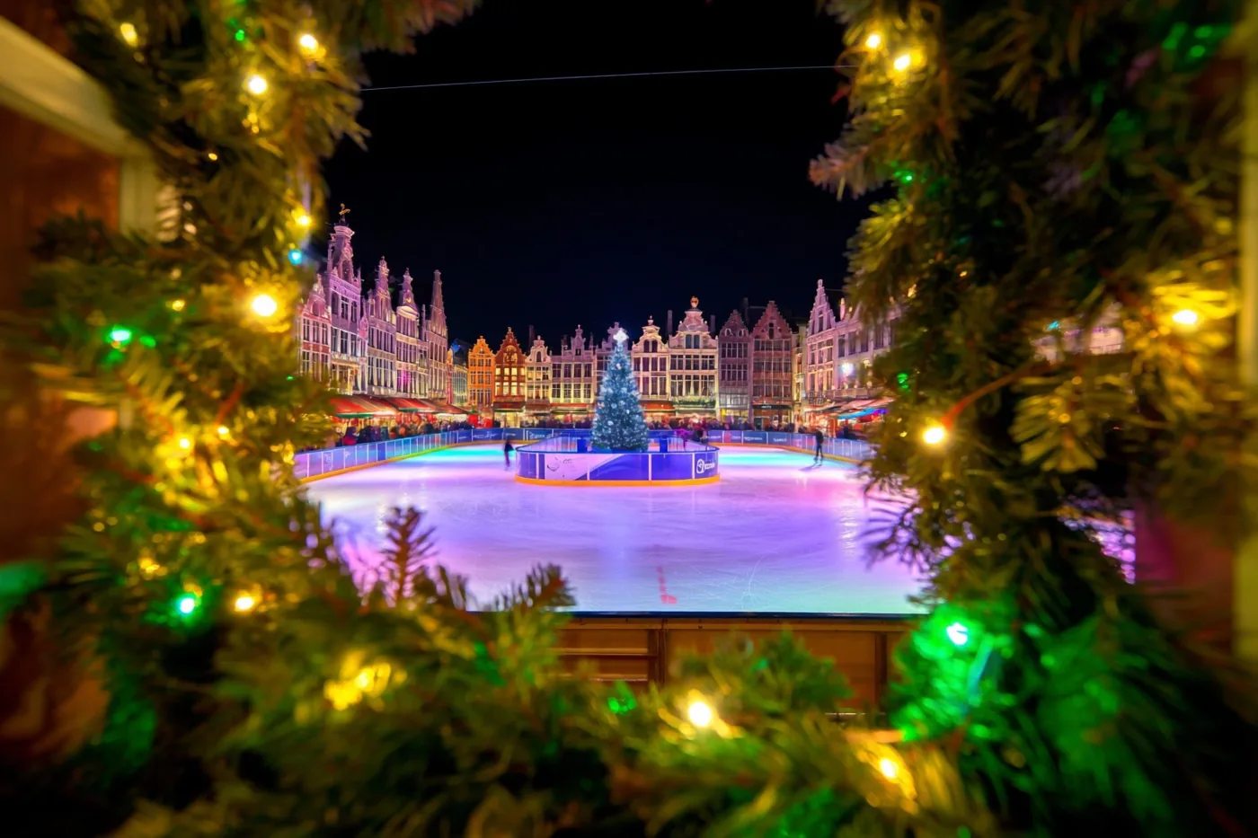 Een nachtelijke ijsbaan op een Belgisch marktplein wordt omlijst door versierde groenblijvende takken met lichtjes, met kleurrijke weerspiegelingen op het ijs, een verlichte kerstboom en schaatsers voor historische gebouwen.