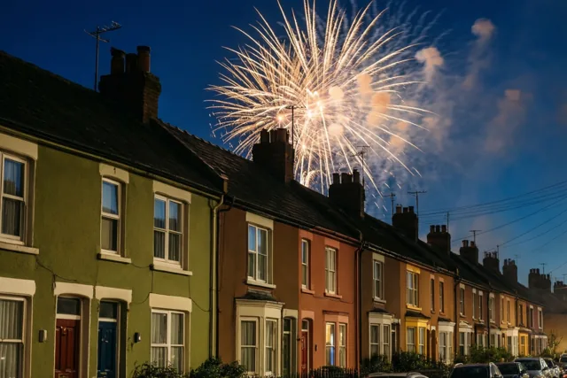 Wit vuurwerk barst los achter de daken van kleurrijke rijhuizen, met warm verlichte ramen onder een donkerblauwe nachtlucht.