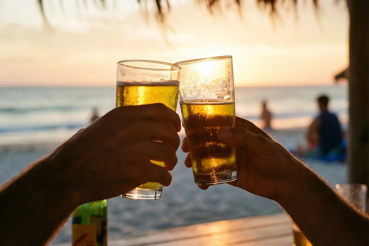 Twee handen klinken met glazen bier bij een strandbar tijdens zonsondergang, in warme gouden en oranje tinten.