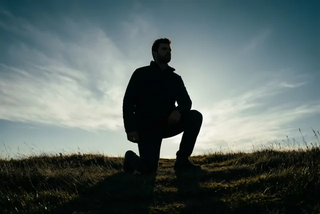 Vanuit een laag standpunt is een man te zien die op één knie knielt op een grasheuvel, als silhouet tegen een heldere hemel met dunne wolken.