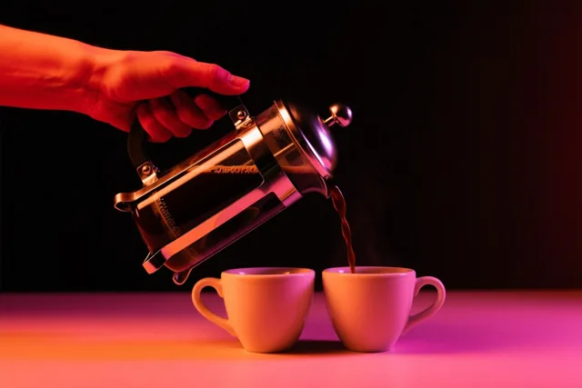 Een hand schenkt koffie uit een cafetière in twee witte kopjes, met rode en roze belichting tegen een zwarte achtergrond.