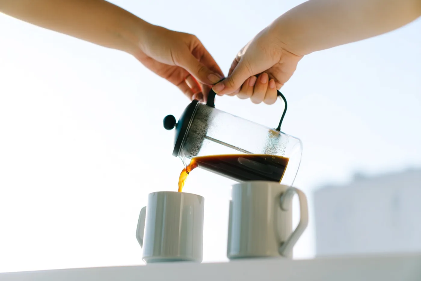 Twee handen schenken donkere koffie uit een glazen cafetière in twee witte keramieken mokken op een witte ondergrond, tegen een lichte, wazige achtergrond.