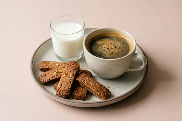 Een bord met een grote kop koffie, een klein glas melk en rechthoekige speculooskoekjes staat tegen een effen lichtroze achtergrond.