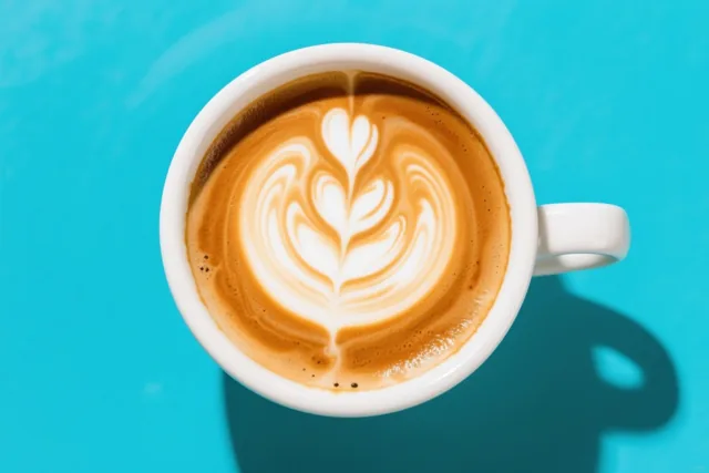 Een witte keramieken kop koffie met een blad- of hartvormig melkschuimpatroon staat op een fel turquoise achtergrond en werpt een zachte schaduw.