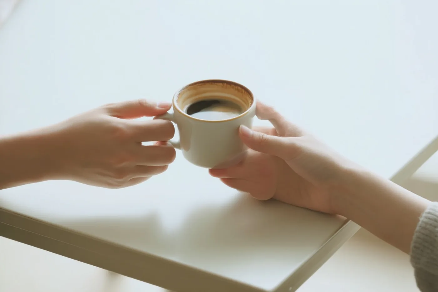 Twee handen houden samen een klein keramieken koffiekopje vast boven een witte tafel tegen een lichte, sobere achtergrond.