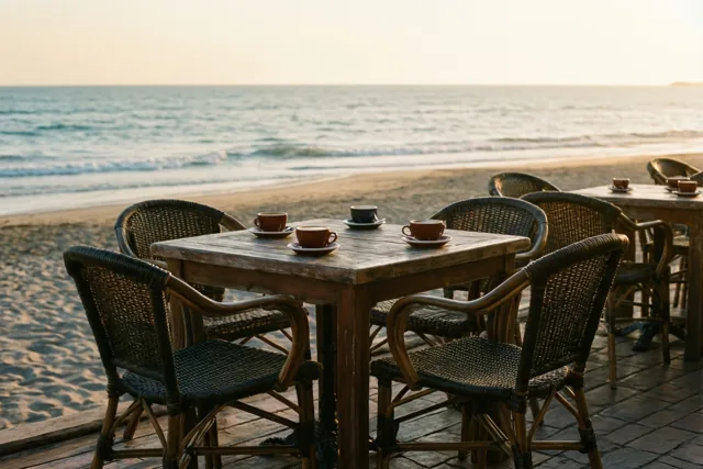 Op een strandterras staan een houten tafel met koffiekopjes en rieten stoelen in goudkleurig ochtendlicht, met uitzicht op zee.