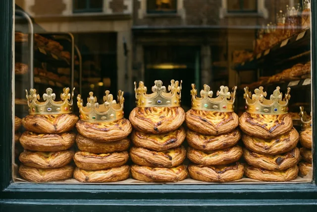 In een warm verlichte bakkersetalage met een donkergroen raamkozijn liggen stapels goudbruine koningentaarten, elk met een gouden papieren kroon.