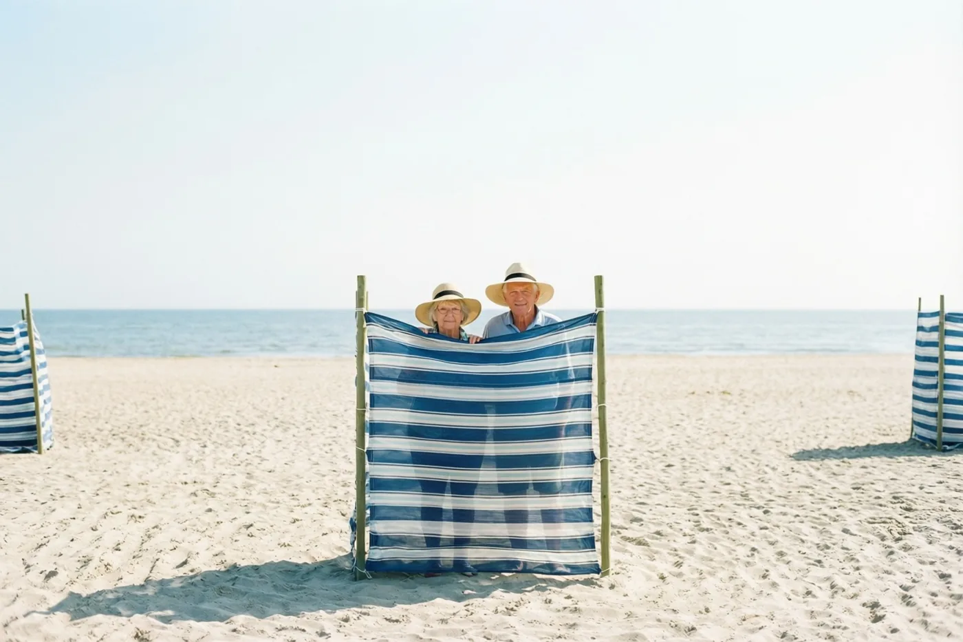 Een ouder koppel met zonnehoeden gluurt van achter een blauwwit gestreept windscherm op een zonnig zandstrand, met een kalme zee en een heldere hemel erachter.