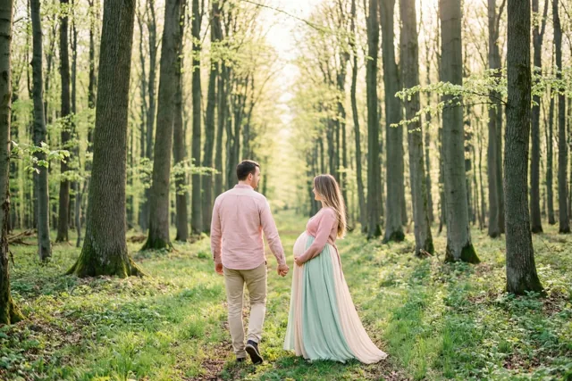 Een zwangere vrouw in een pastelkleurige jurk en een man in een roze hemd wandelen hand in hand door een zonnig beukenbos, met zijn andere hand op haar buik.