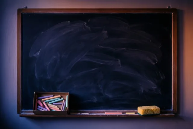 Een afgeveegd krijtbord met een houten kader hangt in paars en oranje licht, met krijtvegen op het bord en een doos gekleurde krijtjes en een gele spons op de richel.