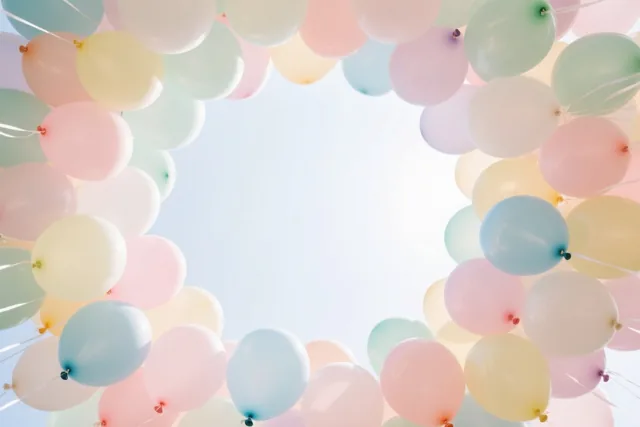 Van onderaf gezien vormen zachtroze, lichtblauwe en lichtgele ballonnen een cirkel tegen een heldere hemel.