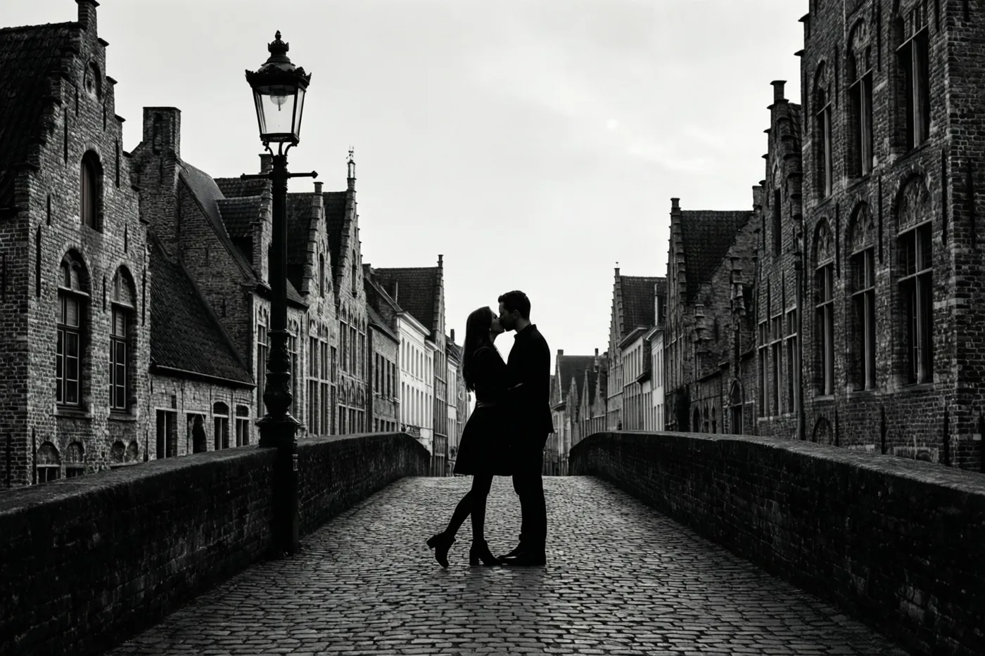 Een zwart-witfoto toont de silhouetten van een kussend koppel op een kasseien brug tussen historische Vlaamse bakstenen gebouwen en een oude straatlamp.