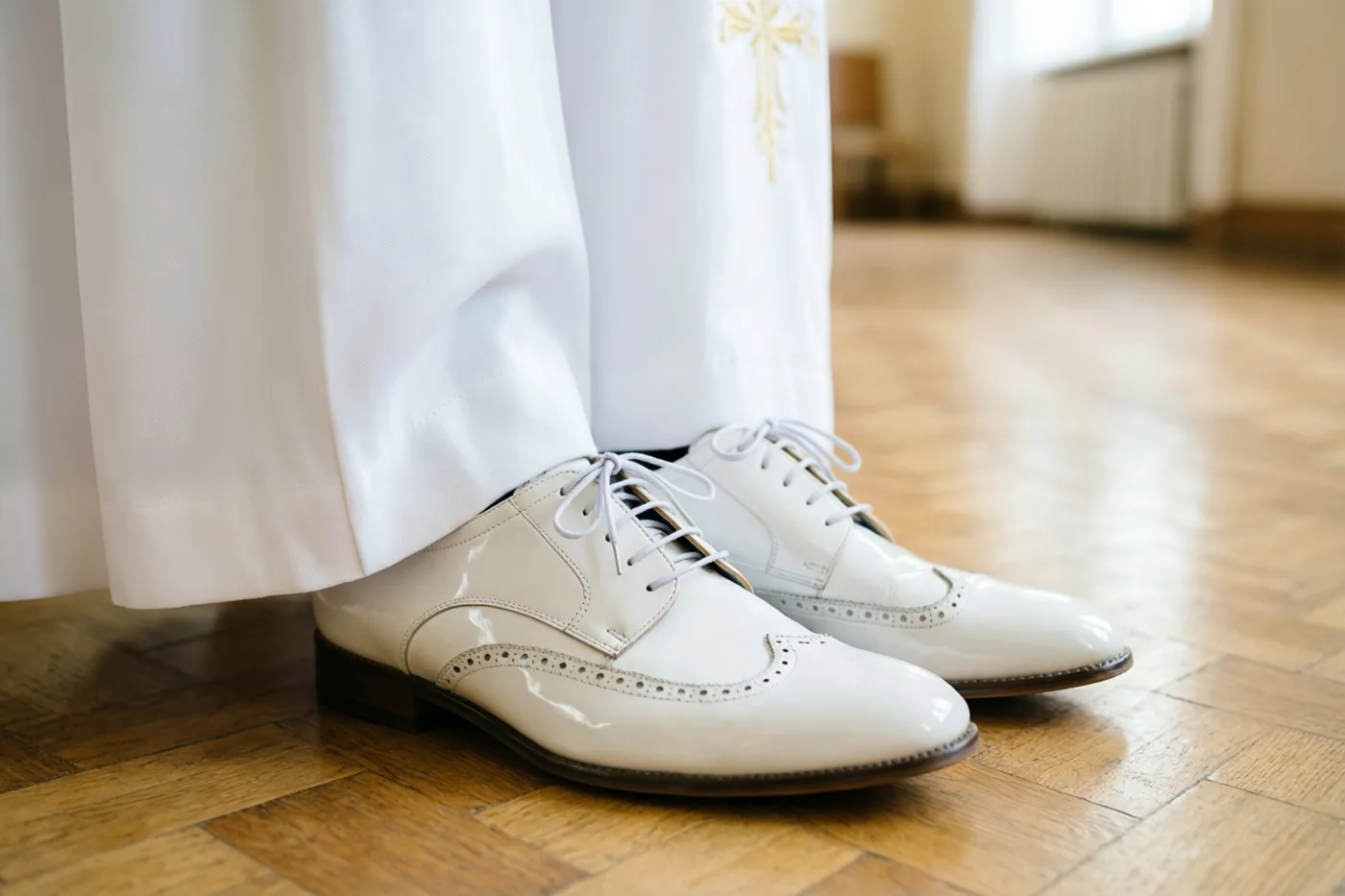 Een paar witte lakschoenen met gaatjesversiering staat op een houten parketvloer, gedeeltelijk bedekt door de zoom van een wit communiegewaad.
