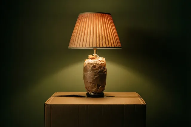 Een in bruin papier gewikkelde lamp brandt op een kartonnen verhuisdoos voor een donkergroene muur.