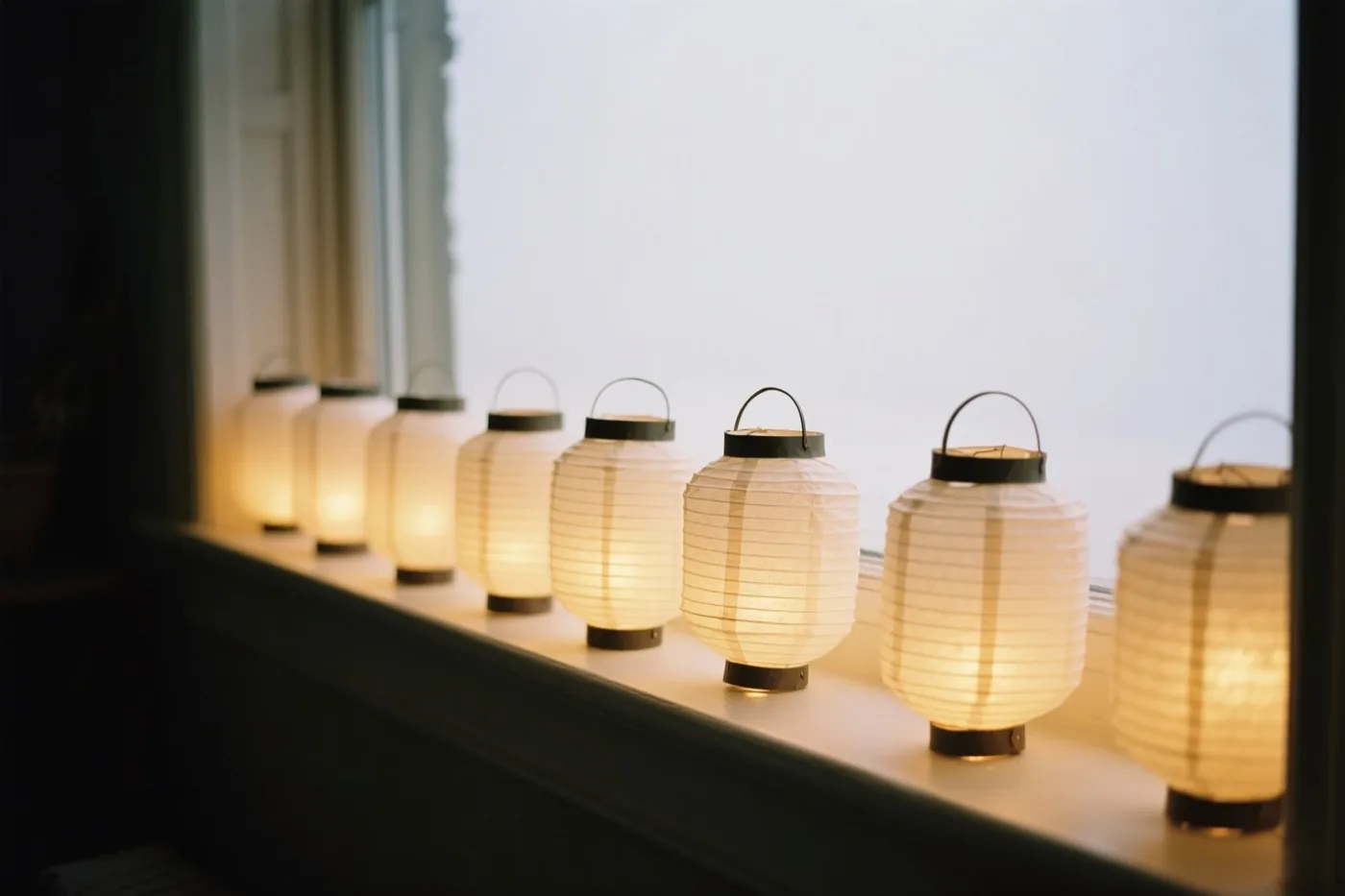Een rij brandende papieren lampionnen staat op een vensterbank met een helder, onscherp raam op de achtergrond.