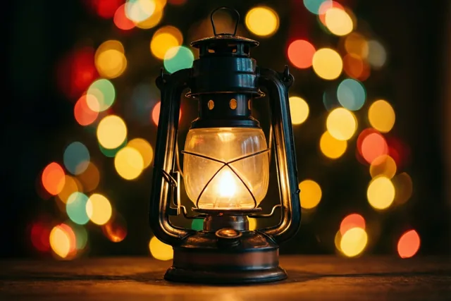 Een brandende lantaarn staat scherp op de voorgrond, met onscherpe feestlichtjes in warme gele, rode en groene tinten erachter.