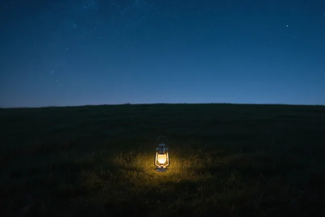 Een lantaarn verspreidt geel licht in een donkere weide onder een sterrenhemel in diepblauwe en zwarte tinten.