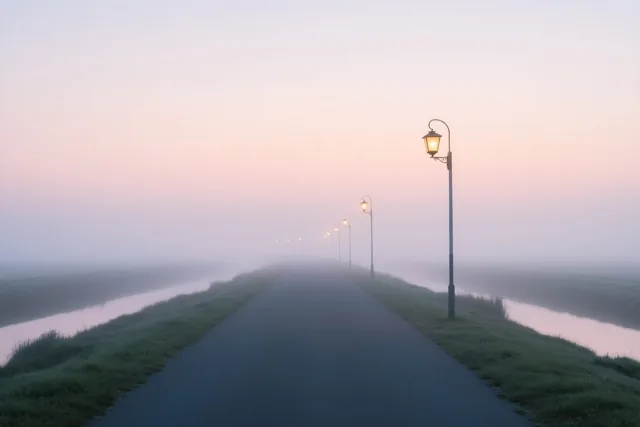 Een mistige polderweg met ouderwetse brandende lantaarns loopt tussen grasbermen en waterlopen naar de verte, onder een roze en lila schemerlucht.