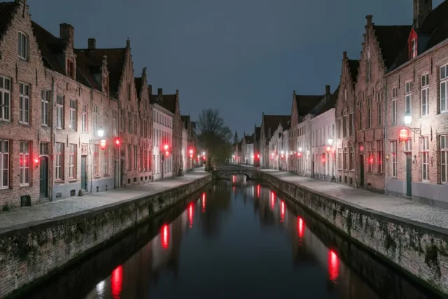 Rode lantaarns verlichten historische bakstenen gebouwen langs een stadsgracht in een Vlaams stadje en weerspiegelen in het water onder een donkerblauwe nachthemel.