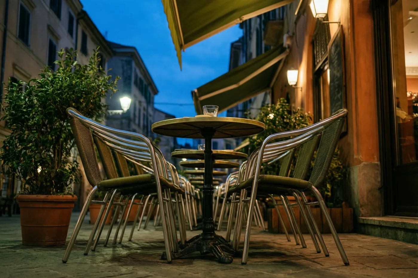 Een leeg caféterras met rijen stoelen en tafels ligt in de schemering voor gebouwen onder een diepblauwe lucht, warm verlicht door straatlichten en licht bij de luifels.
