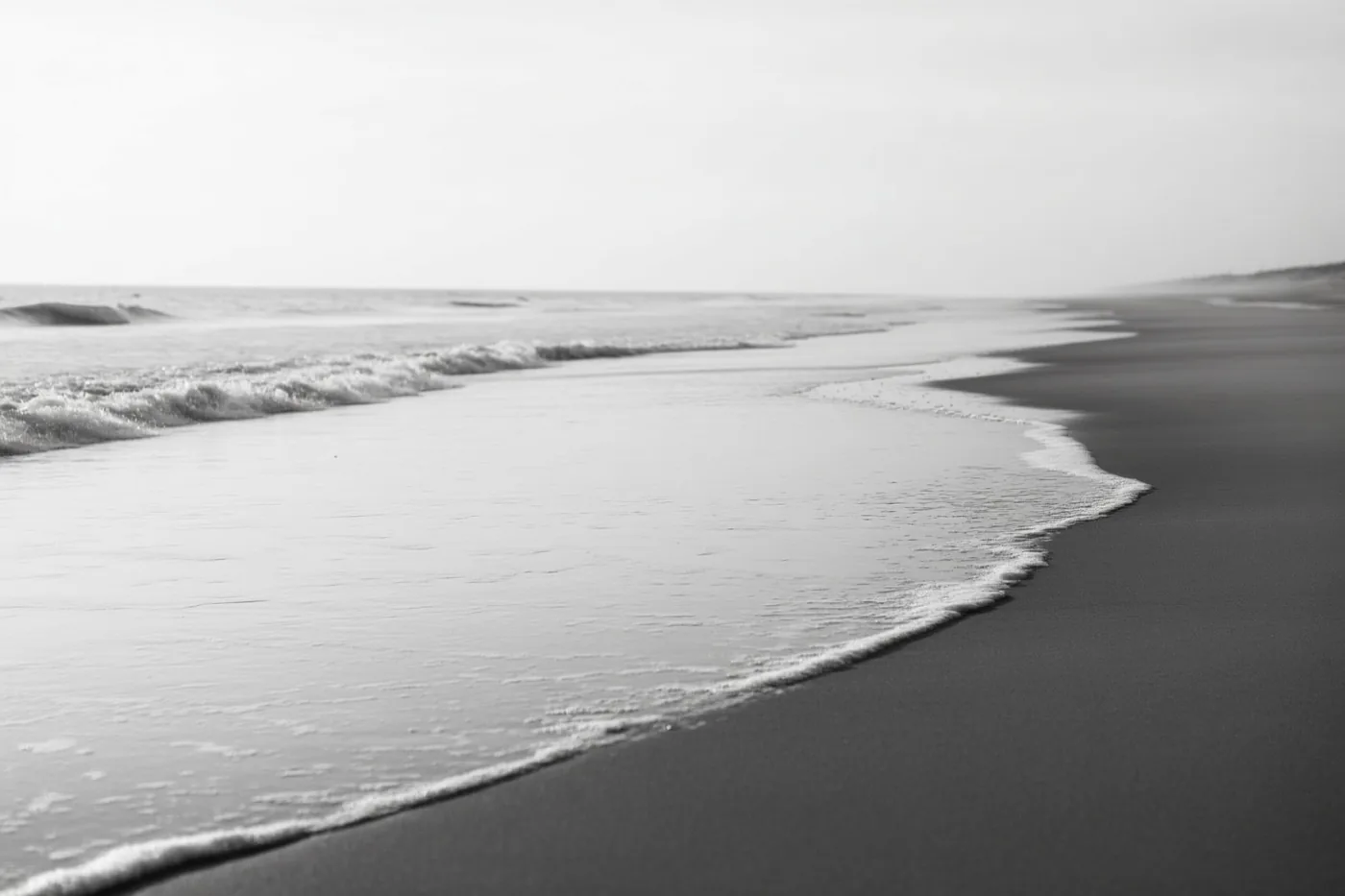 Een zwart-witfoto toont zachte golven op een leeg zandstrand onder een bewolkte lucht.
