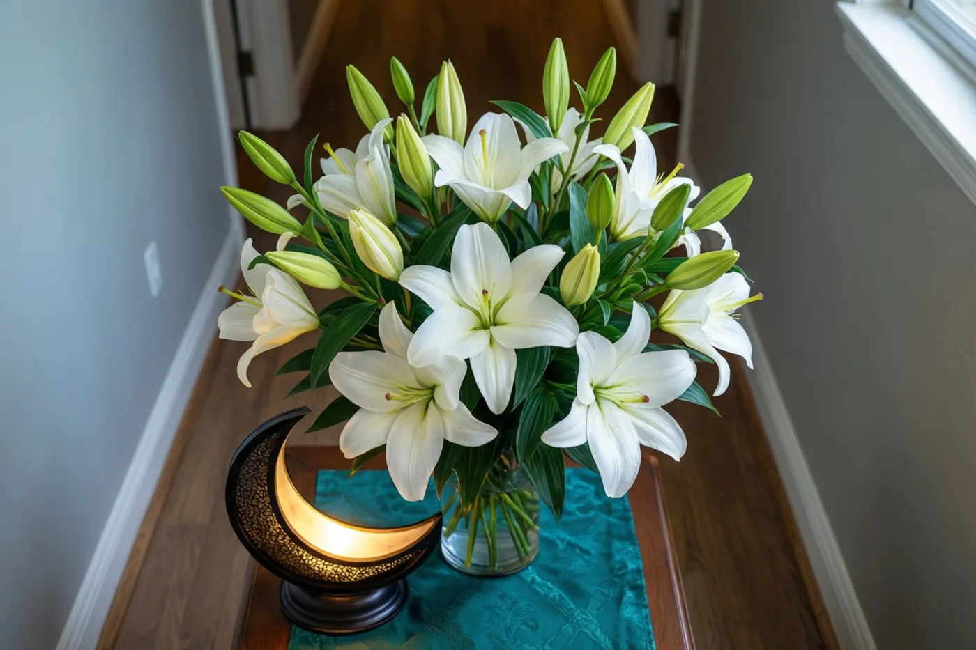 Witte lelies met groene knoppen en bladeren staan in een glazen vaas op een houten tafel met een blauwgroen kleed, naast een lichtgevende maanlantaarn met een donker metalen frame in een gang.