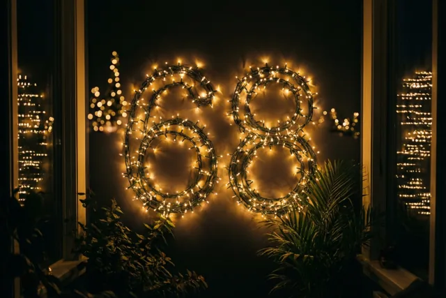 Door een raam is 's nachts het getal 68 in warme lichtslingers op een donkere muur te zien, met planten en onscherpe feestlichtjes eromheen.